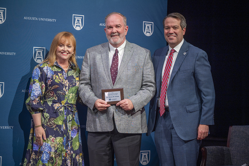 A woman and two men stand in front of an Augusta University backdrop. The man in the middle is holding an award.