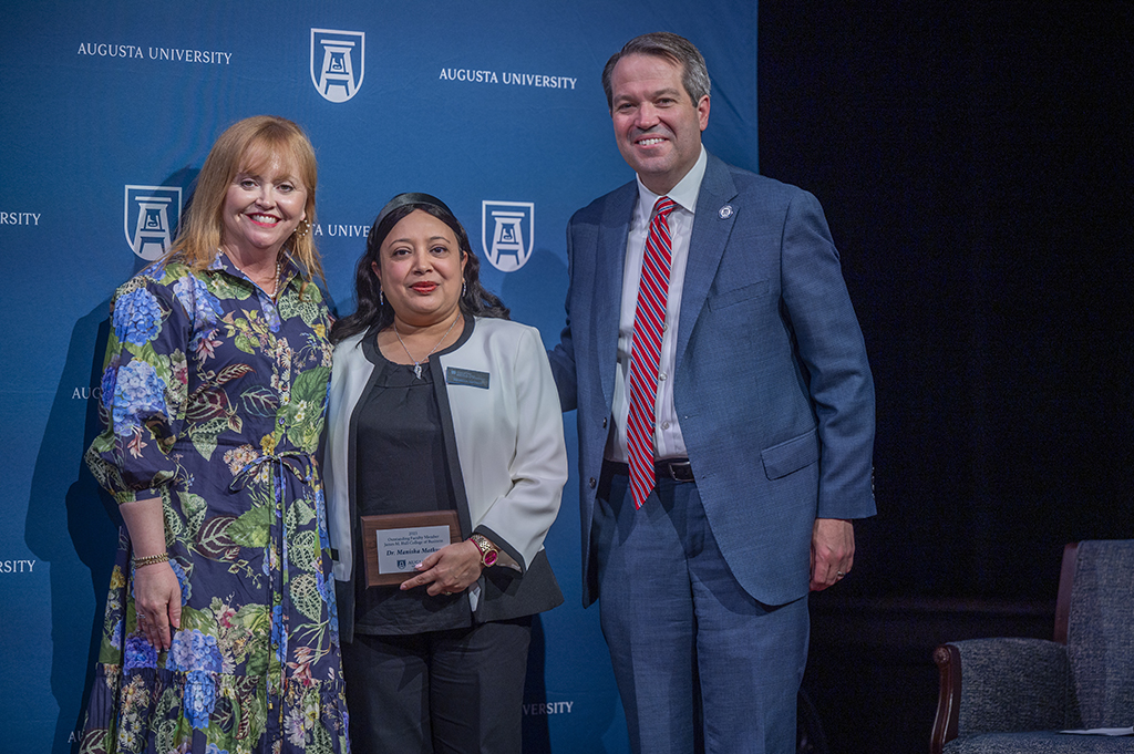 Two women and a man stand in front of an Augusta University backdrop. The woman in the middle is holding an award.