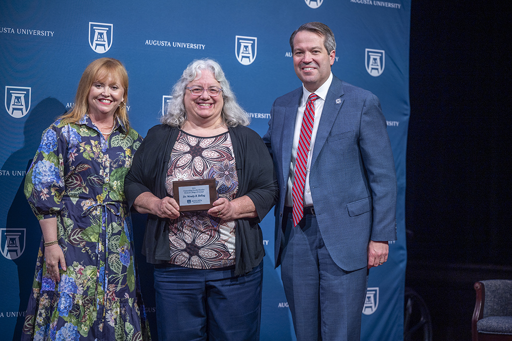 Two women and a man stand in front of an Augusta University backdrop. The woman in the middle is holding an award.