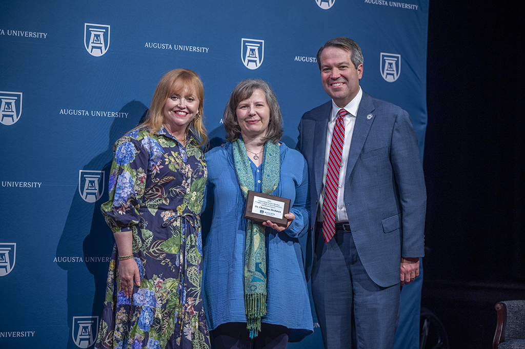 Two women and a man stand in front of an Augusta University backdrop. The woman in the middle is holding an award.