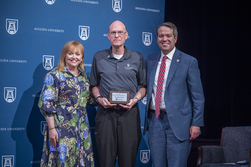 A woman and two men stand in front of an Augusta University backdrop. The man in the middle is holding an award.