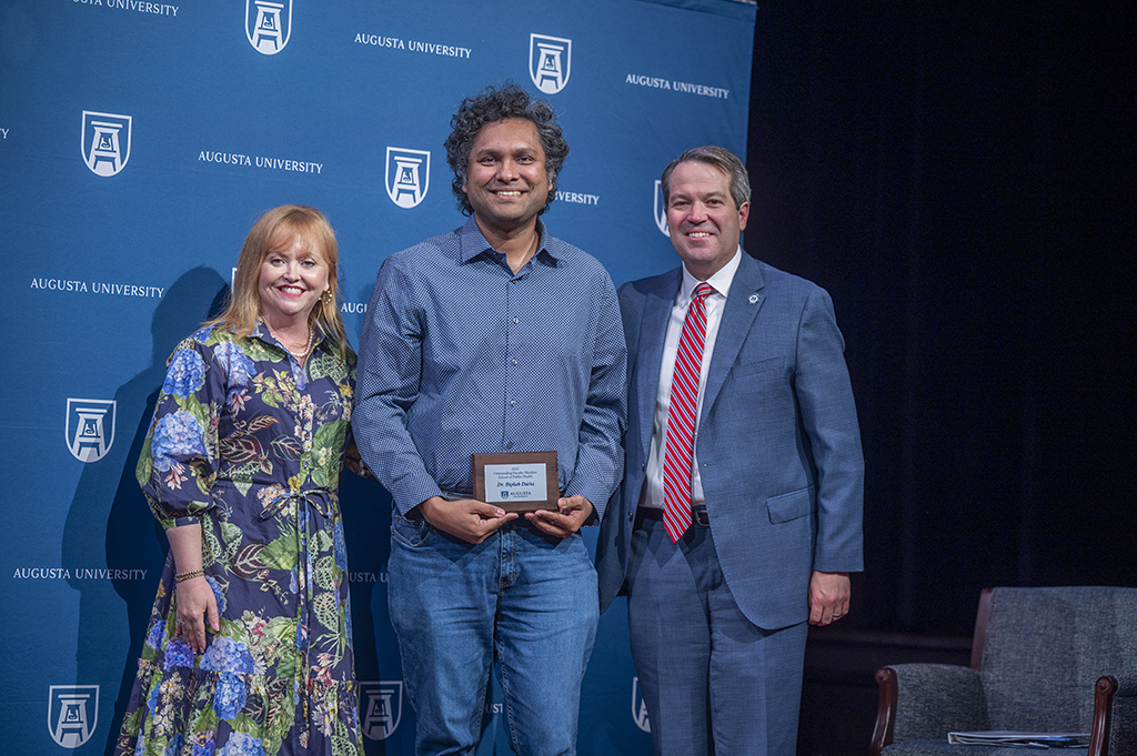 A woman and two men stand in front of an Augusta University backdrop. The man in the middle is holding an award.