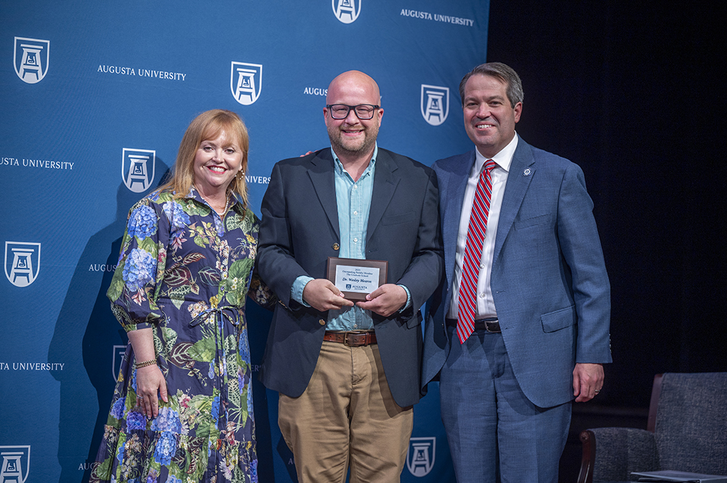 A woman and two men stand in front of an Augusta University backdrop. The man in the middle is holding an award.