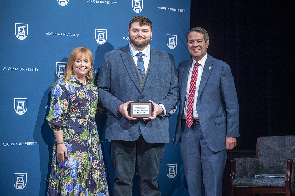 A woman and two men stand in front of an Augusta University backdrop. The man in the middle is holding an award.