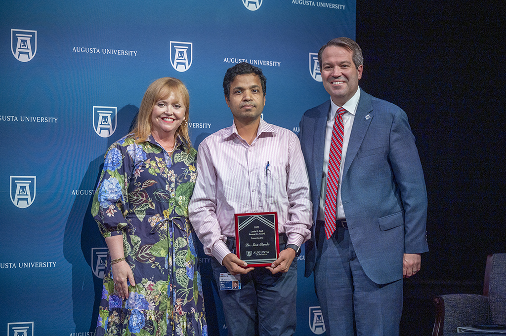 A woman and two men stand in front of an Augusta University backdrop. The man in the middle is holding an award.