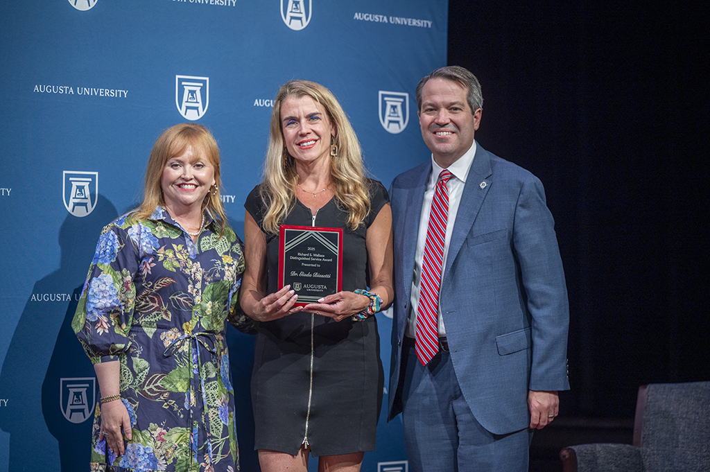 Two women and a man stand in front of an Augusta University backdrop. The woman in the middle is holding an award.