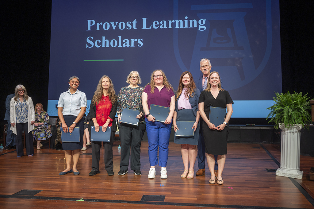 A group of six women and a man stand on a stage holding papers. On the screen behind them says "Provost Learning Scholars."