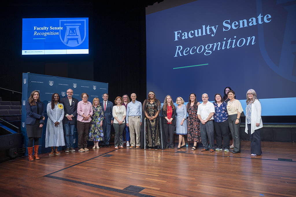 A group of men and women stand on stage. Above and behind them are multiple screens with the words "Faculty Senate Recognition."
