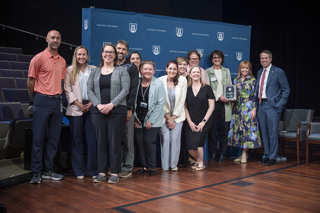 A large group of people pose in front of an Augusta University backdrop. A woman third from right holds an award.