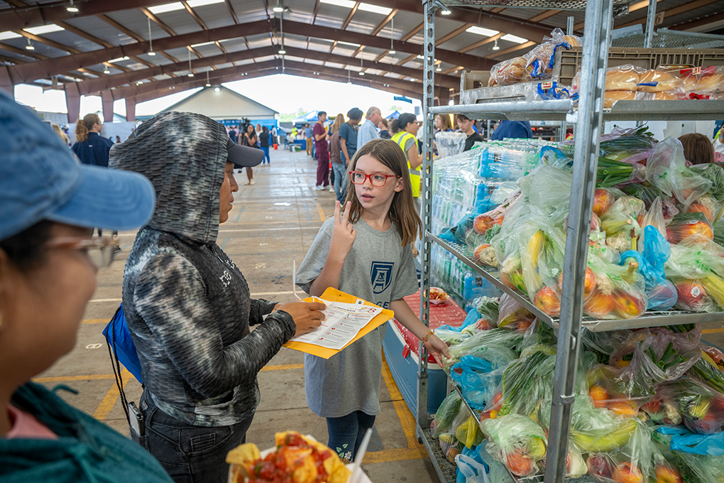 Young girl wearing glasses holds up two fingers to two older women next to fresh food in bags.