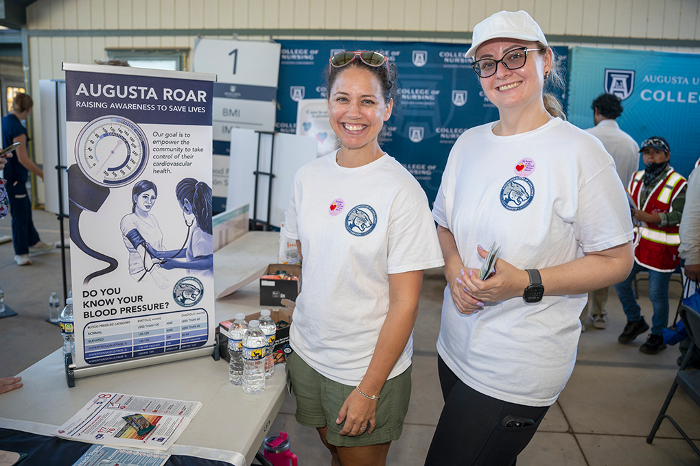 Two women smile in front of a sign holding pamphlets.