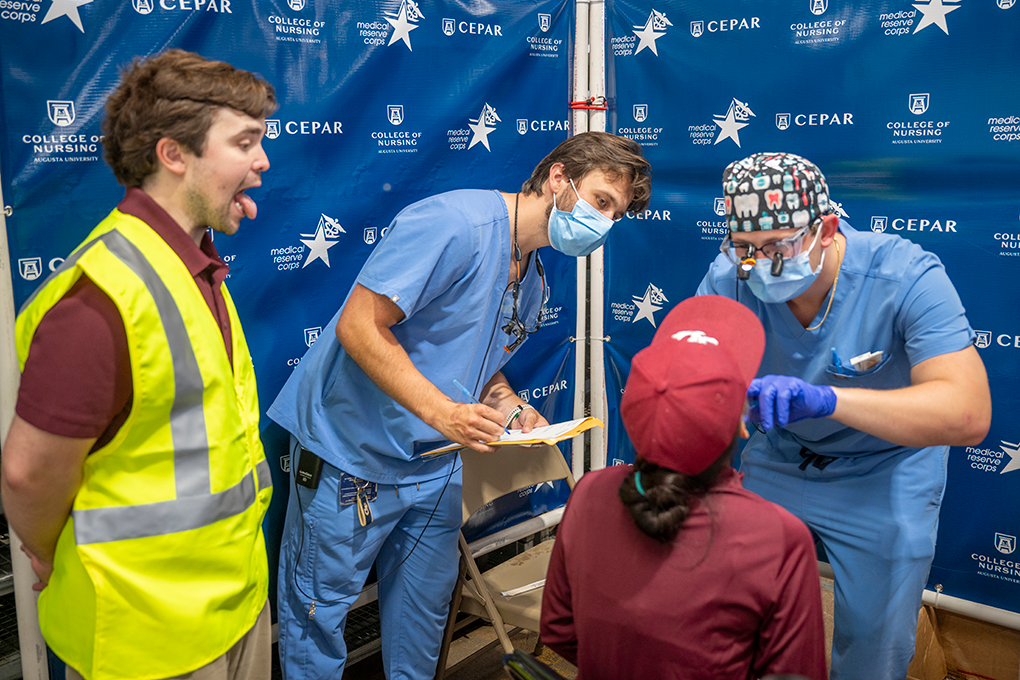 A man in gloves, mask and surgical cap looks inside a woman's throat for a medical exam. Two other young men look on.