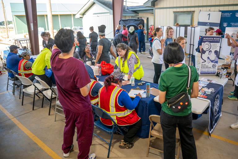 A large crowd of people, some sitting at a table, others standing. Large signage throughout.