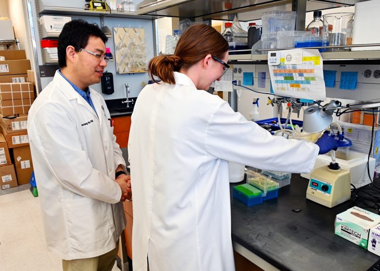 A male scientist and a female high school student work with equipment at a laboratory table.