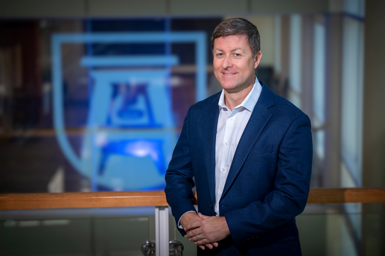 A man in a suit coat leans against a railing on a landing of a set of stairs. Behind him is the logo for Augusta University, which is made up of a bell tower in the shape of an A.