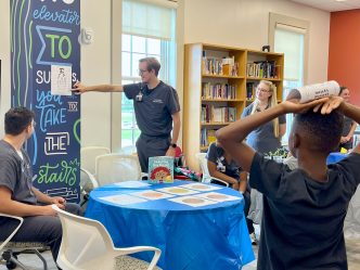 College students studying nursing perform an eye test for a young student inside a modern library. The test is part of a community initiative to help young students bridge the gap between literacy and their health.