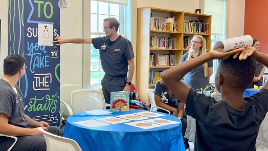 College students studying nursing perform an eye test for a young student inside a modern library. The test is part of a community initiative to help young students bridge the gap between literacy and their health.