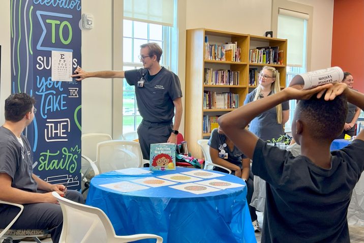 College students studying nursing perform an eye test for a young student inside a modern library. The test is part of a community initiative to help young students bridge the gap between literacy and their health.