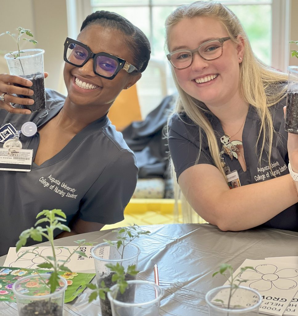 Two college nursing students sit at a table with several tomato plants in plastic cups. The tomato plants were handed out to local grade-school students to encourage healthy eating habits.