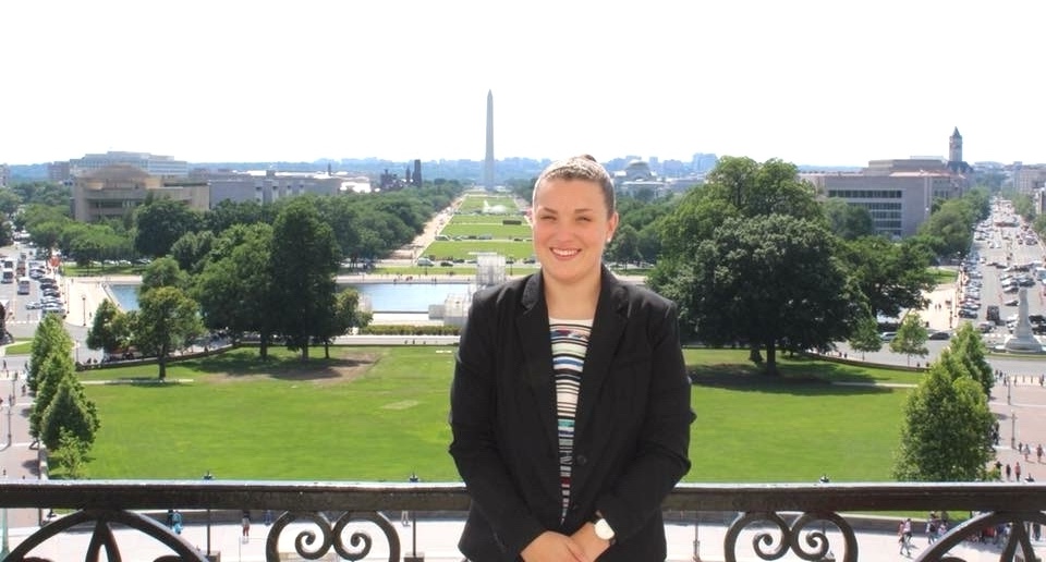 Woman in a black jacket stands on a balcony smiling for the camera with Washington, D.C., in the background.