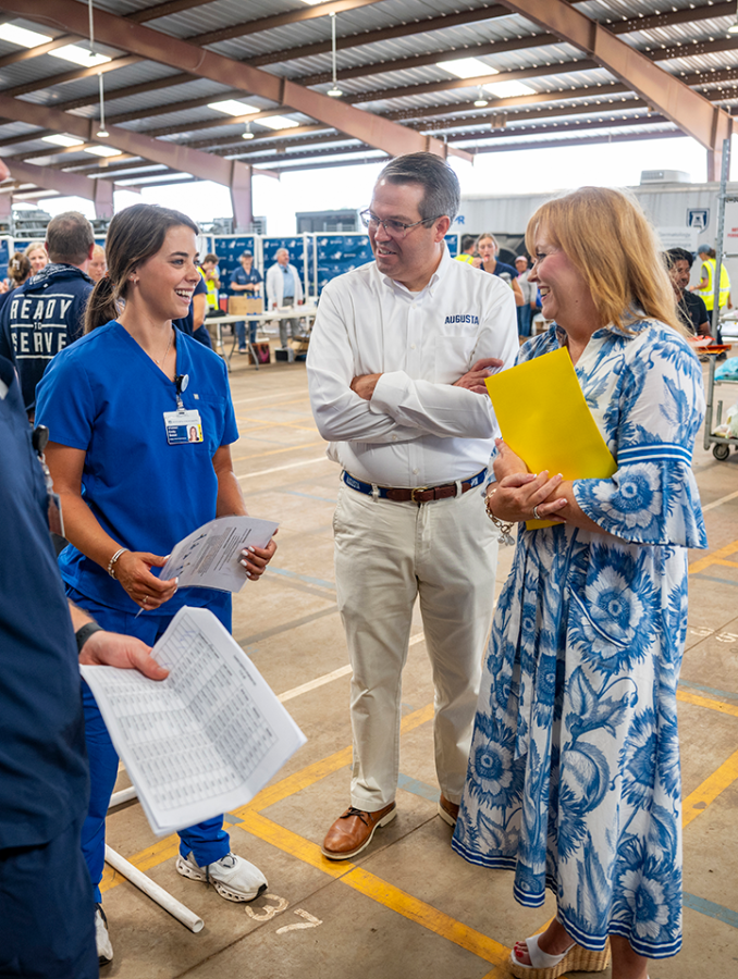 A man with his hands crossed and a woman holding a folder smile standing with a woman in medical scrubs.