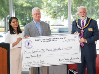 Three people, one woman and two men, stand in front of a large group of people during a check presentation.