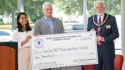 Three people, one woman and two men, stand in front of a large group of people during a check presentation.