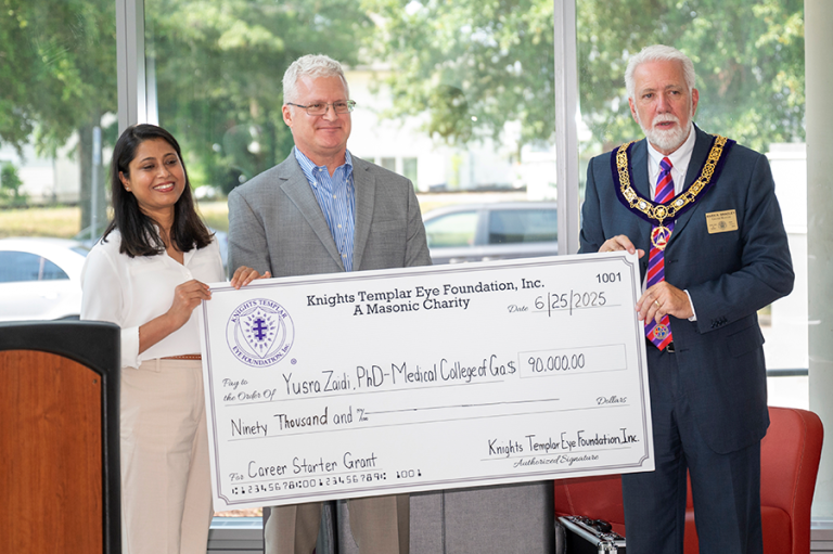 Three people, one woman and two men, stand in front of a large group of people during a check presentation.