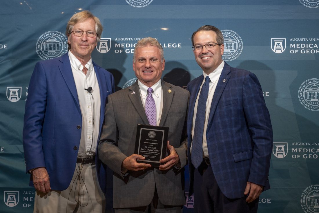 Three men in suits stand in front of a Augusta University Medical College of Georgia backdrop and smile at the camera. The man in the middle is holding a plaque.