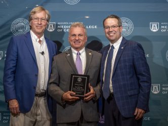 Three men in suits stand in front of a Augusta University Medical College of Georgia backdrop and smile at the camera. The man in the middle is holding a plaque.