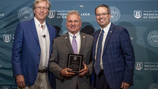 Three men in suits stand in front of a Augusta University Medical College of Georgia backdrop and smile at the camera. The man in the middle is holding a plaque.
