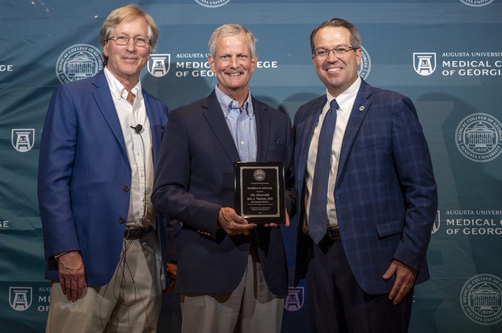 Three men in suits stand in front of a Augusta University Medical College of Georgia backdrop and smile at the camera. The man in the middle is holding a plaque.
