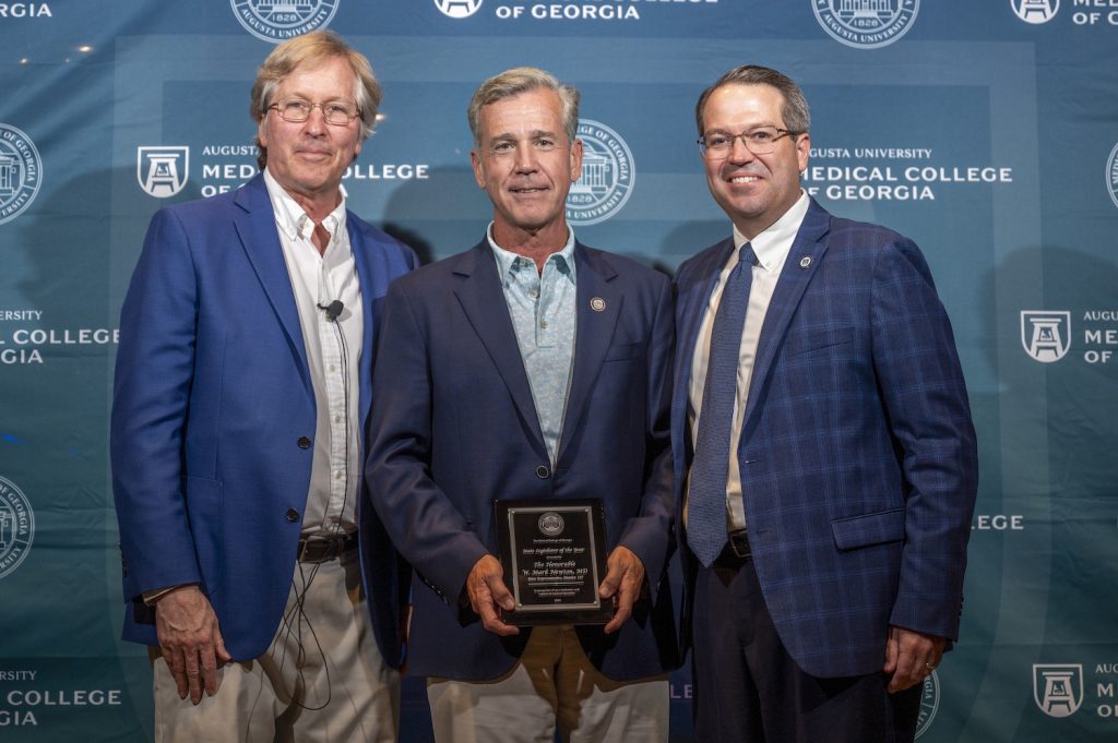 Three men in suits stand in front of a Augusta University Medical College of Georgia backdrop and smile at the camera. The man in the middle is holding a plaque.