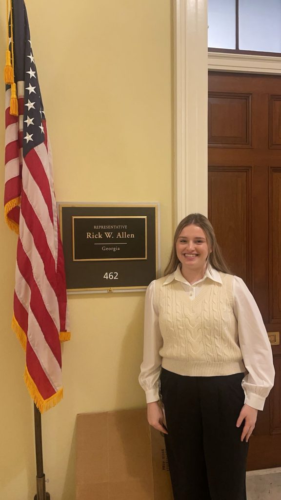 Woman stands in the U.S. Capitol Building next to an American flag and a sign that says "Representative Rick W. Allen, Georgia, 462."