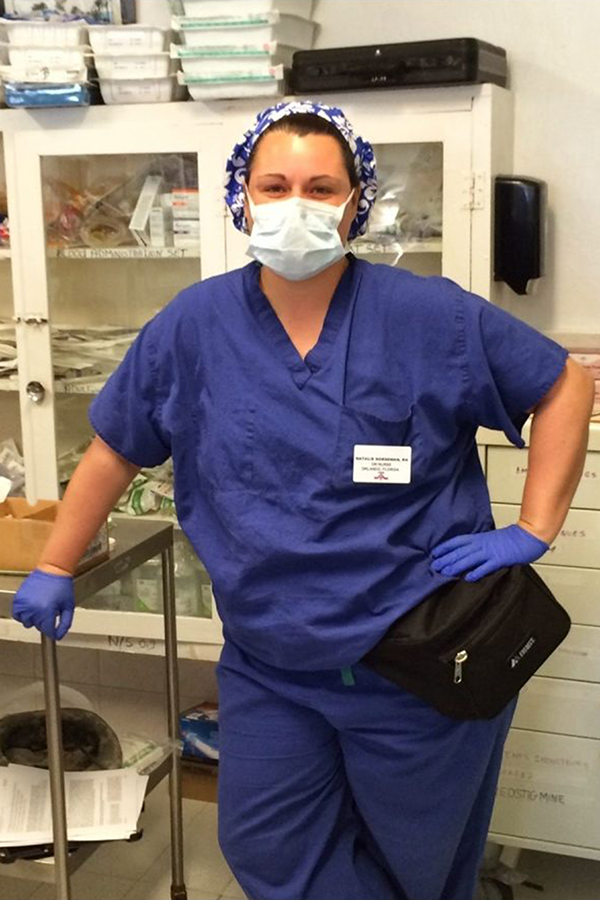 Woman in scrubs, scrub cap, mask, and gloves poses in front of medical supplies
