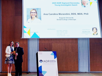 A well-dressed female periodontist and a man stand in front of a projector with the periodontist's face on it. She hold a gold award.