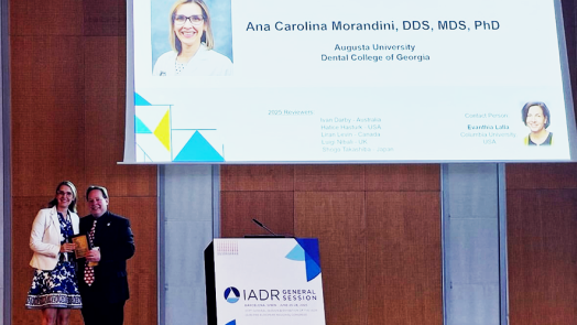 A well-dressed female periodontist and a man stand in front of a projector with the periodontist's face on it. She hold a gold award.