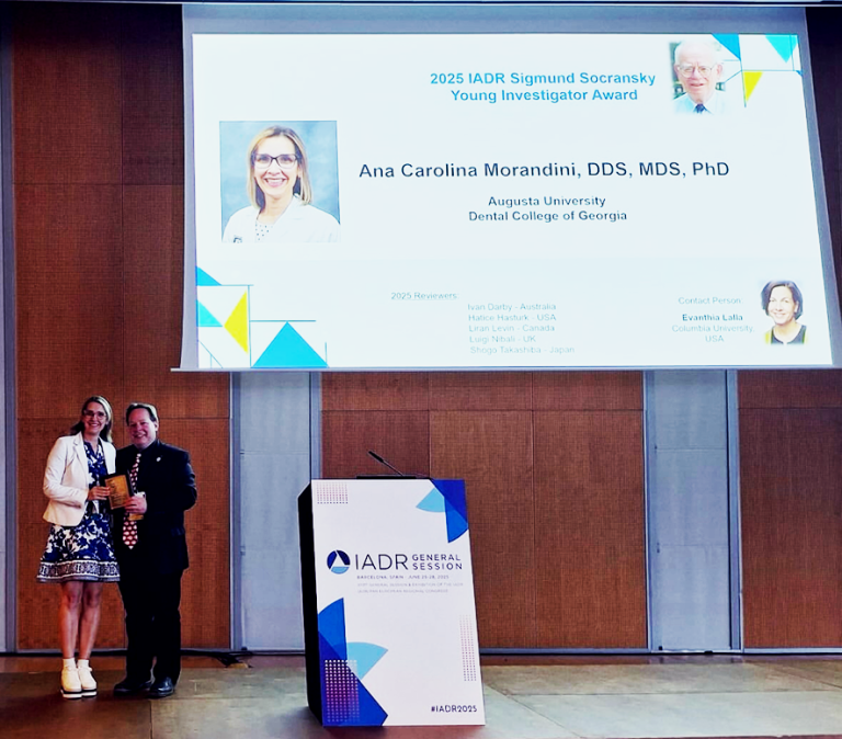 A well-dressed female periodontist and a man stand in front of a projector with the periodontist's face on it. She hold a gold award.