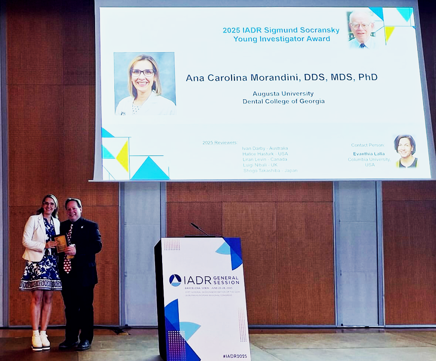 A well-dressed female periodontist and a man stand in front of a projector with the periodontist's face on it. She hold a gold award.