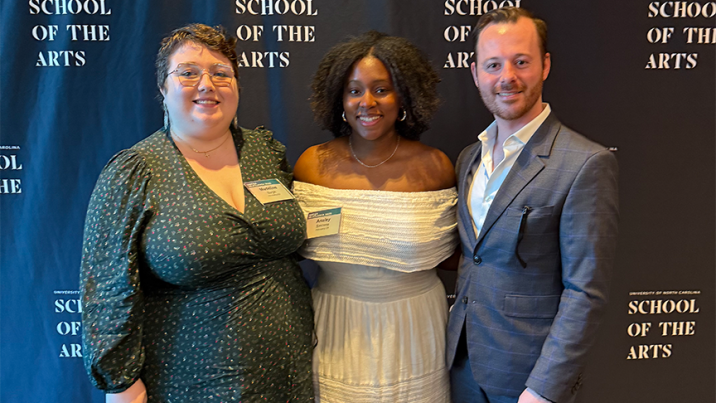 Two women and one man stand together in front of a backgrop during a red carpet event.