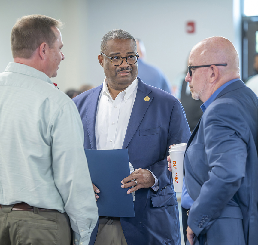 Three men talking. One is holding a folder and one is holding a coffee.