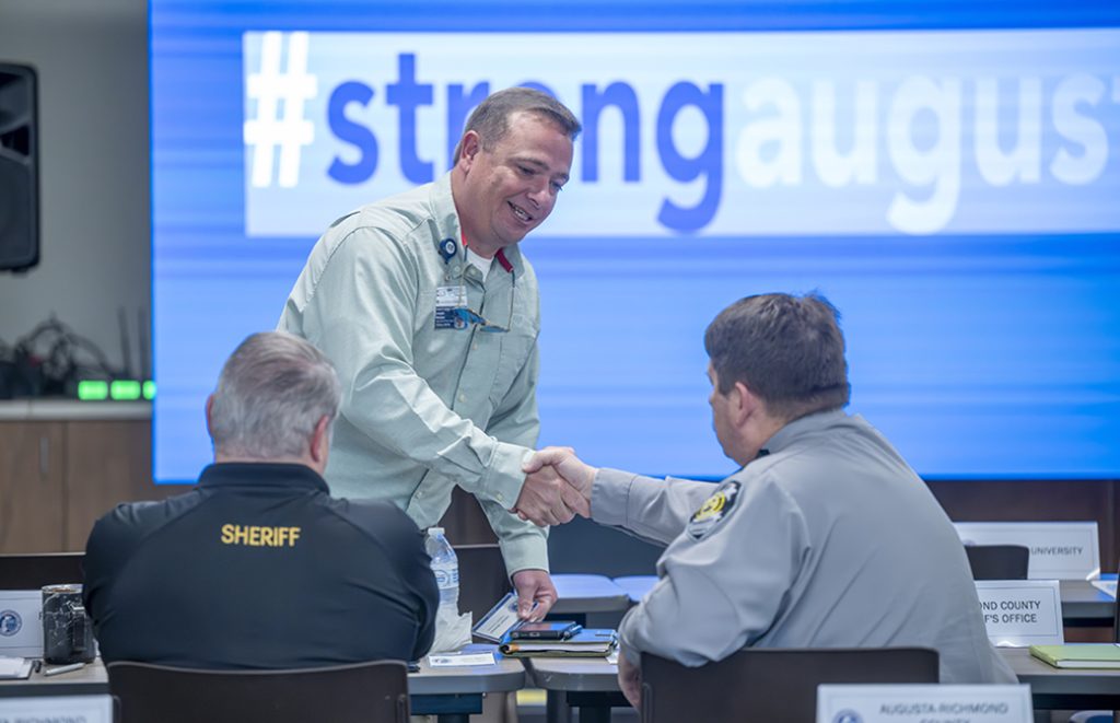 A man standing shakes hands with two men sitting at tables. On the screen in the background is "#strongaugusta"