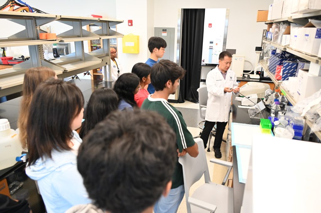 A group of high school and college students tour a scientific lab. A scientist is showing them different equipment and how it works.