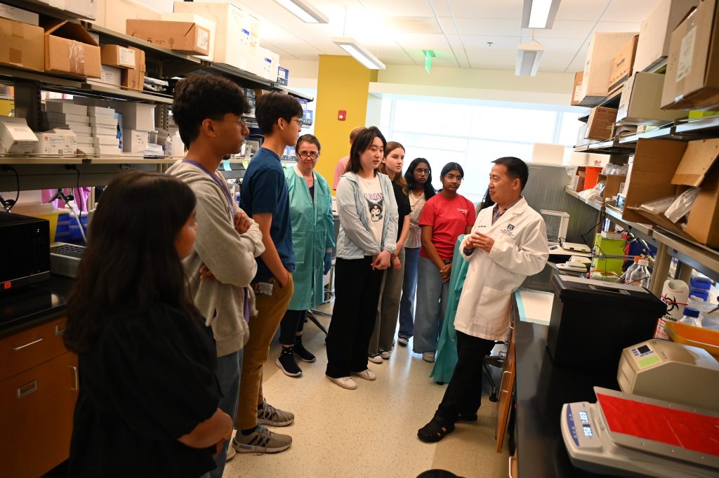 A group of high school and college students tour a scientific lab. A scientist is showing them different equipment and how it works.