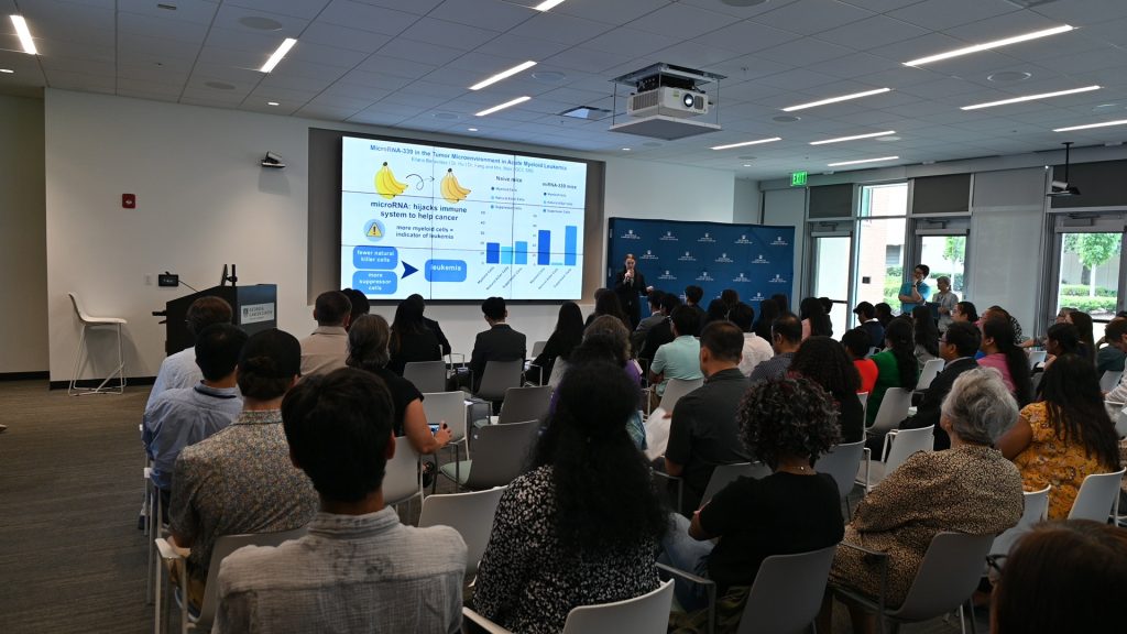 A large group of people sit and listen to high school and college students talking about their experience in a summer scholars program.