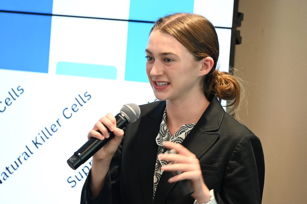 A female high school student gives a presentation in front of a large group. She describes her experience learning about research in a lab.