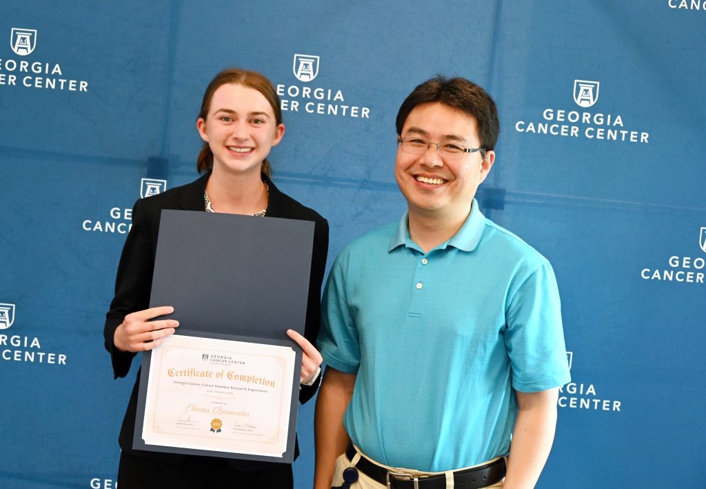 A female high school student stands with a researcher. The high school student is holding up a certificate indicating she completed a summer scholars program.