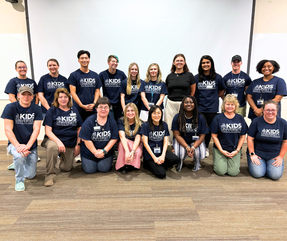A large group of college professors, staff, and students stand in a large classroom. They are all wearing t-shirts that say "Kids University" across the chest.