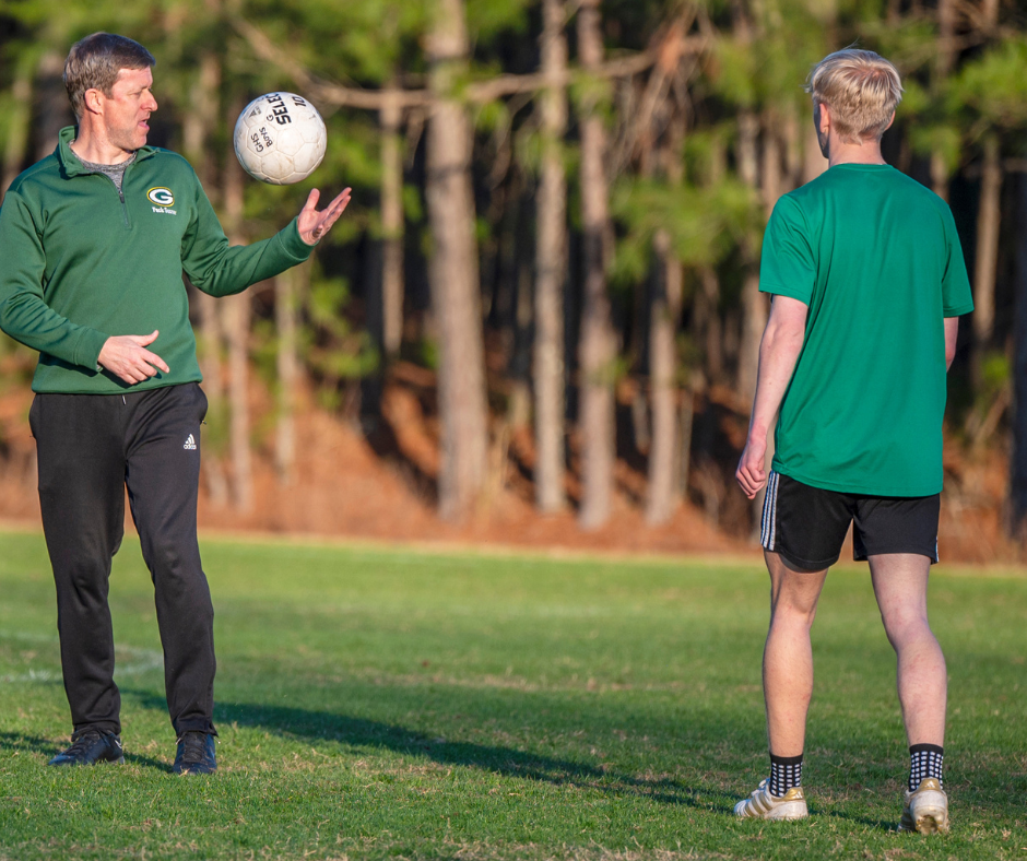 A soccer coach wearing a pullover gestures while tossing a ball during practice, speaking to a student athlete.
