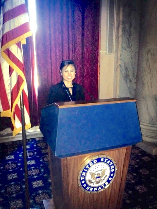 A woman stands at a podium with a seal that says United States Senate on it. Beside her is an American flag.